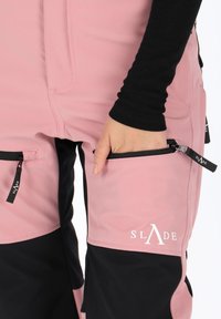 Pink and black ski pants with a smooth exterior, zippered pockets, and logo detailing. Textured fabric, fitted design, and adjustable waistband.