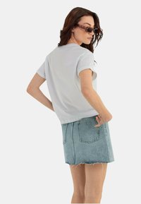 T-shirt bianco a maniche corte abbinato a una gonna di jeans azzurro chiaro. La gonna presenta orlo sfilacciato e tasche posteriori. La modella indossa occhiali da sole.