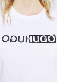 Une personne blonde portant un t-shirt blanc avec un texte noir en gras disant "OGUHUGO" où "HUGO" est mis en avant dans un rectangle noir.