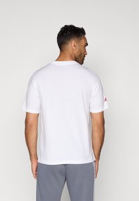 Λευκό βαμβακερό T-shirt με κοντά μανίκια, στρογγυλή λαιμόκοψη και κόκκινη λογότυπη λεπτομέρεια στο αριστερό μανίκι. Φοριέται με γκρι παντελόνι.