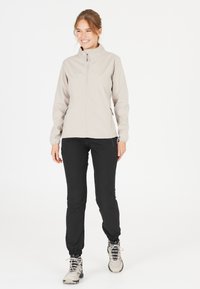 Ljusbeige softshell-jacka med ståkrage, dragkedja fram och svarta byxor, med elastiska ärmslut och en insvängd design.