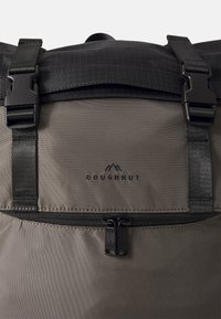 Sac à dos en tissu gris avec un rabat noir, comportant une poche zippée et des bretelles réglables. Logo "DOUGHNUT" embossé sous la fermeture éclair.