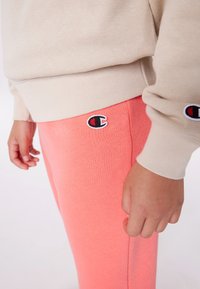 Light beige sweatshirt with a ribbed hem, paired with soft coral joggers featuring an embroidered logo on the thigh. Smooth texture.