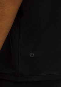 Czarny sportowy T-shirt o gładkiej fakturze, zaokrąglonym dolnym brzegu i małym, okrągłym logo blisko dolnej krawędzi. Widoczne detale szwów.