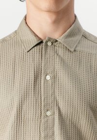 Chemise beige à boutons avec un tissu texturé à motif diamant, un col pointu et une fermeture à boutons standard. Matière douce.
