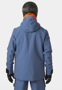 Giacca invernale blu in tessuto con cappuccio, dotata di guanti neri, dettagli arancioni nel colletto e un design elegante e moderno.