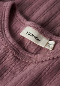 Różowy dzianinowy top z prążkowanym wzorem, z falbaniastym dekoltem oraz metką z logo "Lil'Atelier" i informacjami o rozmiarze. Miękka tekstura z pionowymi liniami.