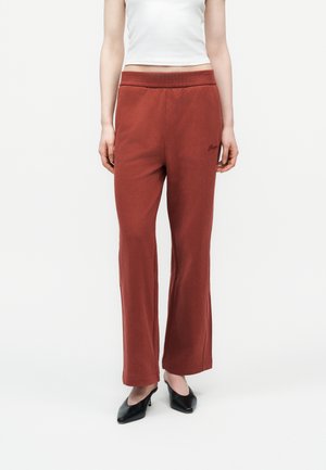 Kobieta w szerokich spodniach w kolorze rdzy z elastycznym pasem i czarnych butach na obcasie, stojąca na białym tle.