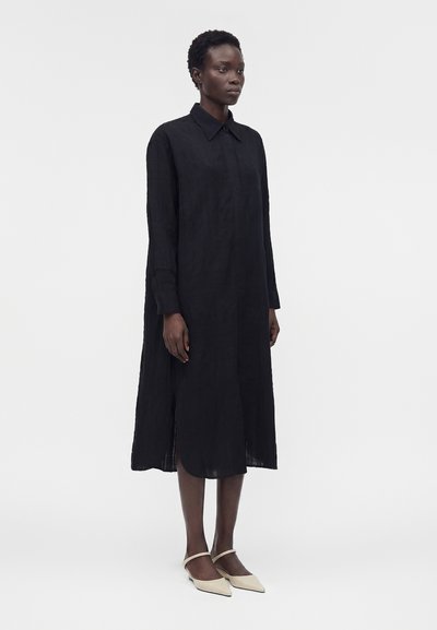 Γυναίκα με κοντά μαλλιά που φοράει μαύρο midi φόρεμα με μακριά μανίκια και μπεζ μπαλαρίνες με μυτερή μύτη, στέκεται μπροστά σε ένα μονόχρωμο ανοιχτό φόντο.