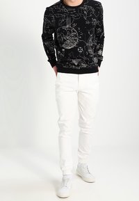 Maglione nero con stampe grafiche bianche, abbinato a pantaloni bianchi e sneakers chiare. Il maglione presenta un collo a giro e polsini a costine.