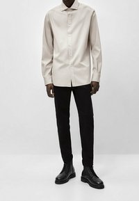 Jasnobeżowa koszula na guziki, czarne obcisłe spodnie oraz czarne sznurowane buty. Koszula ma zaostrzony kołnierzyk i długie rękawy.