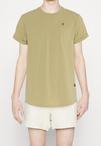 Olivově zelené tričko s koulenými krátkými rukávy, kulatým výstřihem a malým logem na hrudi. V kombinaci se světle krémovými texturovanými šortkami.