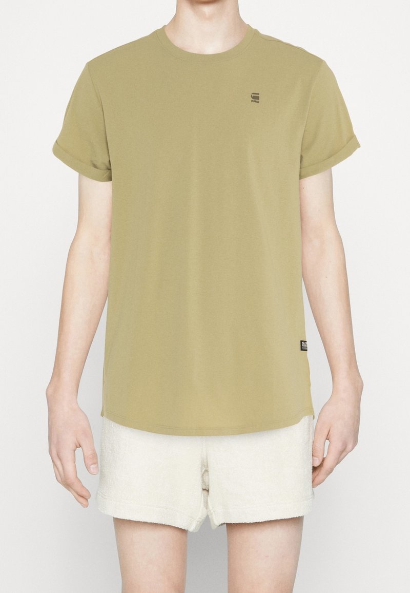 Olivově zelené tričko s koulenými krátkými rukávy, kulatým výstřihem a malým logem na hrudi. V kombinaci se světle krémovými texturovanými šortkami.