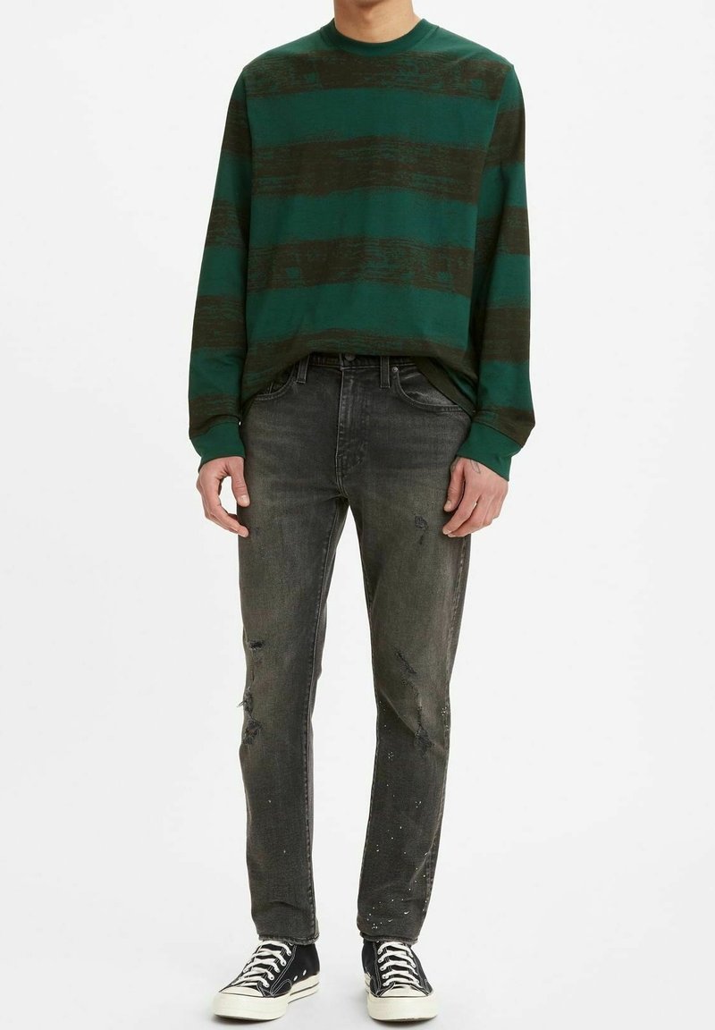 Camisa de manga larga a rayas verdes y negras combinada con jeans oscuros desgastados y zapatillas negras. Material de algodón, corte relajado, crea un look informal.