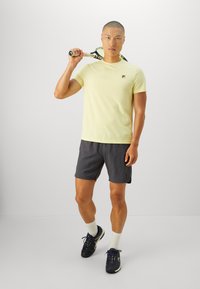 Aktiivne mees kollases T-särgis, tumedates lühikestes pükstes ja spordijalatsites, hoides tennisereketit õlal, seistes ühetaolise taustaga.