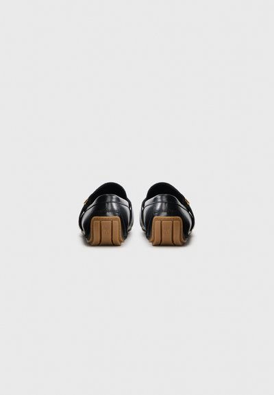 Svarta läderslip-on skor med en slät finish, med en beige gummisula och minimalistisk design. Inga framträdande mönster eller hårdvara synliga.