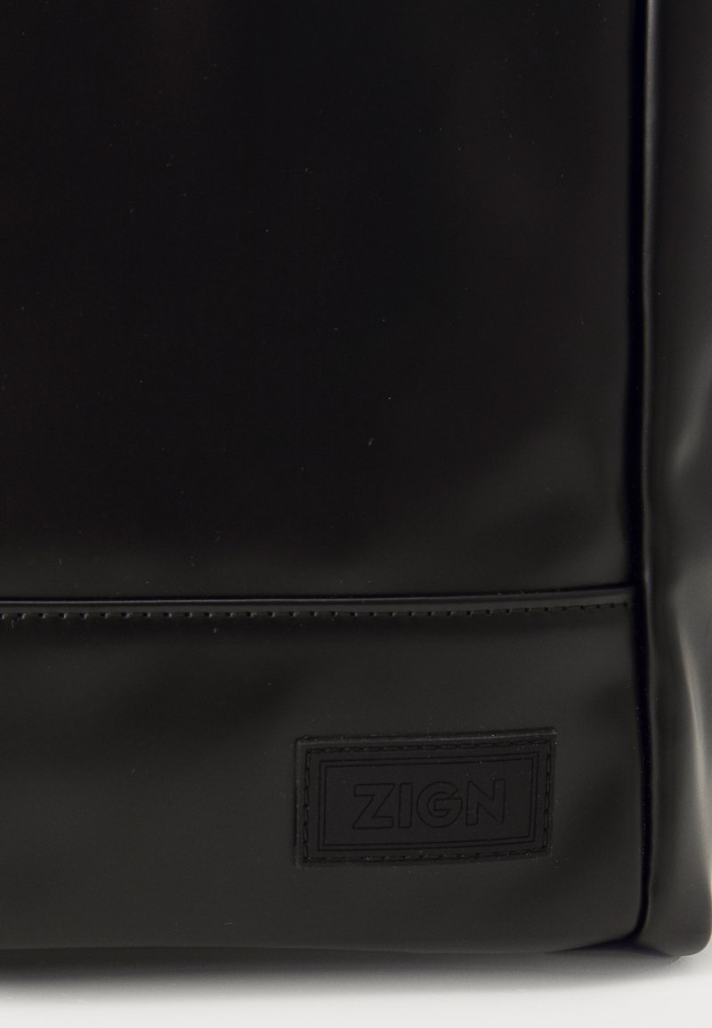 Schwarze, glänzende Tote-Tasche mit glatter Textur, ausgestattet mit einer genähten Naht und dem eingeprägten "ZIGN"-Logo auf einem rechteckigen Patch.