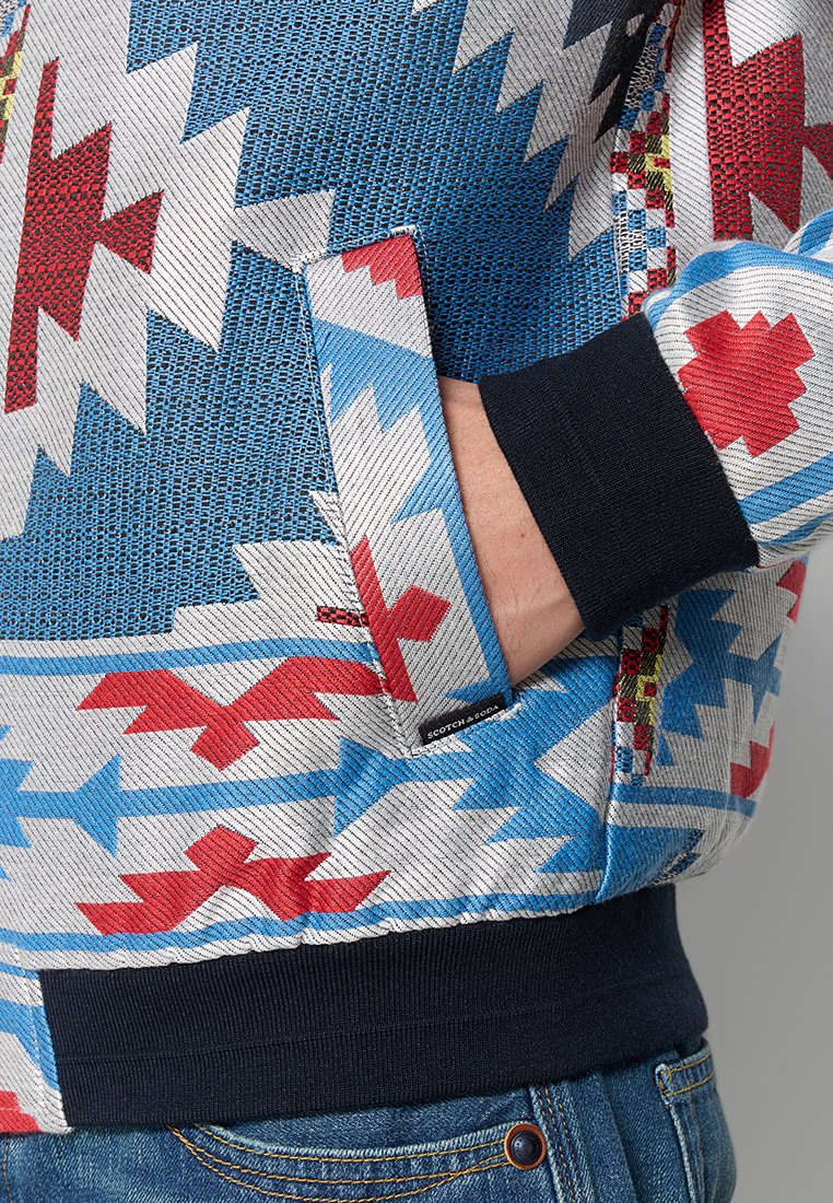 Hand in de zak van een blauw, wit en rood gemusterd jasje met zwarte manchetten, gecombineerd met blauwe spijkerbroek.