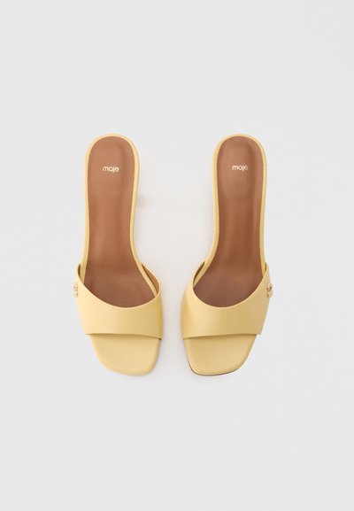 Κίτρινα σανδάλια με λουρί, με λείο δερμάτινο φινίρισμα, διαθέτουν φαρδύ λουρί και μικρό διακοσμητικό κουμπί. Καφέ εσωτερική σόλα με μινιμαλιστικό σχεδιασμό.