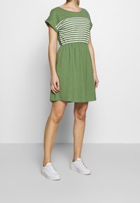 Grünes Kleid mit kurzen Ärmeln, gestreiftem Oberteil, elastischem Bund und ausgestellt Rock. Kombiniert mit weißen Sneakers. Einfaches, lässiges Design.