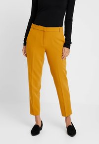 Żółte spersonalizowane spodnie o gładkiej teksturze, wąskim kroju i szlufkach na pasek, zestawione z czarnymi mokasynami i dopasowaną czarną bluzką.