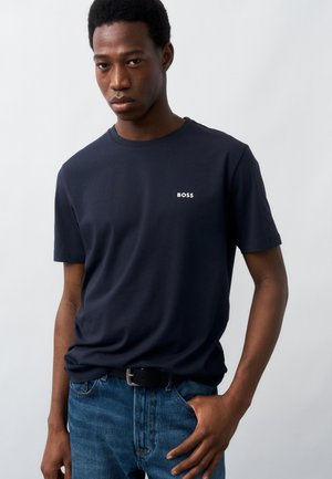 Νέος άνδρας με μπλε σκούρο t-shirt BOSS και μπλε τζιν, ποζάρει με το ένα χέρι στην τσέπη μπροστά σε απλό φόντο.