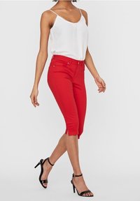 Röda capribyxor med figurnära design, utrustade med midjebyte och slitsar på sidorna vid fållen, kombinerade med en vit camisole och svarta klackskor.