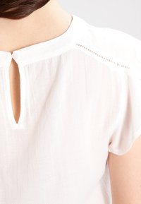Blusa blanca de manga corta, con una pequeña apertura en forma de ojal en la parte posterior del cuello y una sutil textura en la tela.