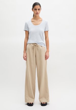 Vrouw met donker haar in een paardenstaart, gekleed in een lichtblauw T-shirt met korte mouwen, beige wijde broek met trekkoord en zwarte platformsandalen.