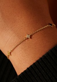 Goldene Gliederarmband mit sternförmigen Gliedern, eines davon mit kleinen klaren Steinen verziert. Die Oberfläche ist glänzend und glatt, getragen am Handgelenk.