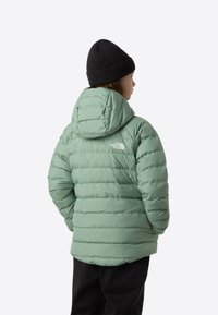 Hele roheline puffer-jakk horisontaalse tikandiga, kapuutsiga ja logoga seljal. Sobitatud musta kootud mütsiga.