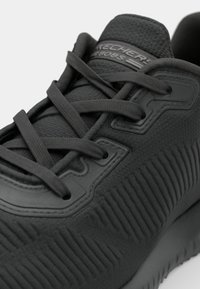 Musta urheilukenkä, jossa on teksturoitu kangaspäällinen, paksu pohja ja tasaiset yhteensopivat nauhat; logo prässättynä kieliosaan. Erityinen poimuttuva kuviointi.