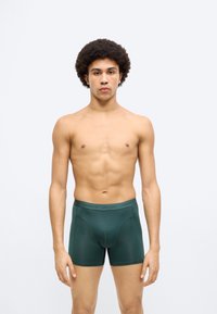 Σκούρα πράσινα μποξεράκια από απαλό ύφασμα, με εφαρμοστή σχεδίαση και μαλακή μέση. Το μοντέλο στέκεται σε ουδέτερη στάση.