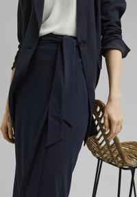 Mörkblå skräddarsydd blazer över en ljus topp, i kombination med en mörkblå omslagkjol med en knutdetalj vid midjan. Vävd stol i bakgrunden.