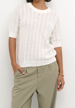 Femme portant un haut en maille blanc à manches courtes avec un motif œillet en chevron et un pantalon taille haute vert olive, main dans la poche, bracelet en or.