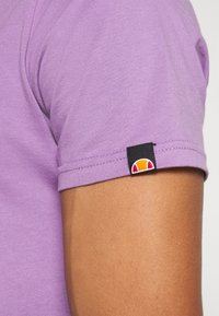 Lila bomull t-shirt med kort ärm, med en väl sydd nederkant. En svart etikett med en flerfärgad logotyp syns på ärmen.