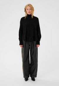 Moteris su garbanotais šviesiais plaukais vilki juodą įgulos kaklą turintį megztinį ir plačius juodus odinius kelnes, stovinti ant balto fono.