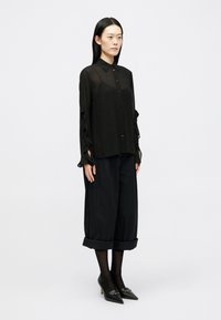 Camisa preta transparente com detalhes em godé nas mangas, combinada com calças largas cropped pretas e sapatos de salto agudo pretos. Textura suave.