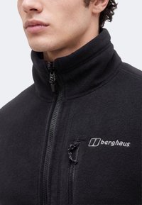 Κοντινό πλάνο ατόμου που φοράει μαύρο fleece μπουφάν Berghaus με φερμουάρ στο ψηλό γιακά και τσέπη στο στήθος.