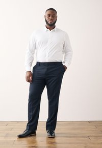 Weißes Langarmhemd, marineblaue Hose und schwarze Schuhe. Das Hemd hat einen Knopfkragen; die Hose hat einen flachen Vorderteil und seitliche Taschen.