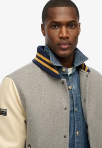 Chaqueta tipo varsity de mezcla de lana gris con mangas de cuero crema y cuello azul marino con rayas amarillas, superpuesta sobre una camisa de denim.