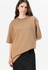 Smeđa oversized majica kratkih rukava s okruglim izrezom, s toniranim Adidas logotipom sprijeda, u kombinaciji s crnom suknjom.
