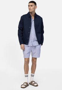 Άνδρας που φοράει ναυτικό μπουφάν πάνω από μπλε ριγωτό πουκάμισο και σορτς, λευκές κάλτσες με μαύρες ρίγες και καφέ σανδάλια, στέκεται μπροστά σε λευκό φόντο.