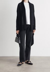 Svart öppen stickad cardigan över en vit topp, ihop med svarta raka jeans och svarta flata sandaler med rosettaccent. Liten svart handväska.