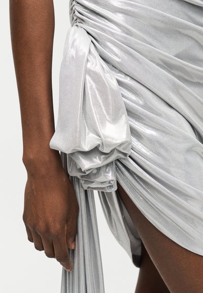 Άτομο με σκουρόχρωμο δέρμα φοράει λαμπερό ασημένιο ρούχο μαζεμένο που δένεται σε κόμπο στο ισχίο, αποκαλύπτοντας μέρος του γυμνού μηρού και του αριστερού χεριού.