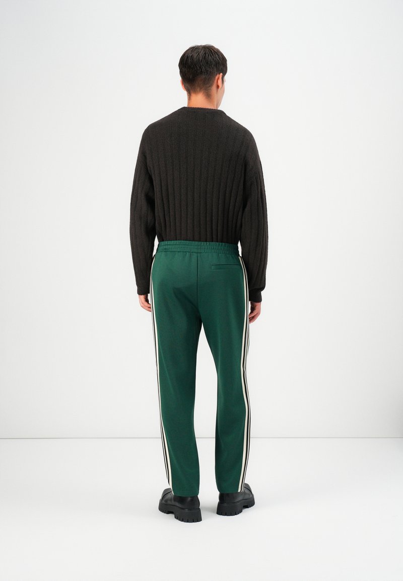 Pull en fausse maille noir et pantalon de survêtement vert avec des bandes blanches sur le côté, doté d'une taille élastique, et associé à des chaussures noires imposantes.