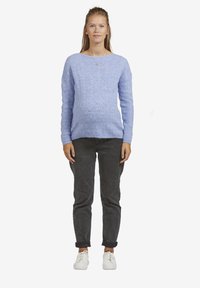 Junge Frau steht mit einem hellblauen Strickpullover, dunkelgerollten Jeans und weißen Sneakers vor einem weißen Hintergrund.