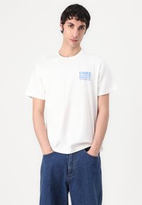 White cotton T-shirt with a blue printed logo on the left chest. The model pairs it with blue denim shorts, hands in pockets.