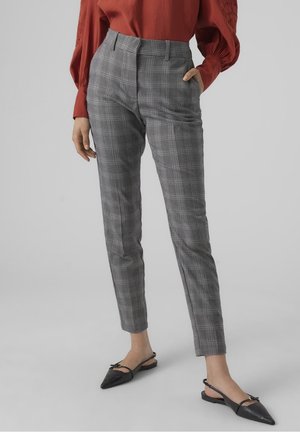 Woman wearing grey checkered tapered pants, red long-sleeve blouse, and black pointed slingback flats, standing with one hand in pocket.