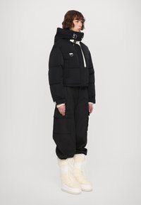 Melna īsā pūkaina jaka ar baltām akcentiem, ar rāvējslēdzēju un auklas aizdari, saskaņota ar brīvām melnām biksēm un krēmkrāsas kažokādas oderes zābakiem.